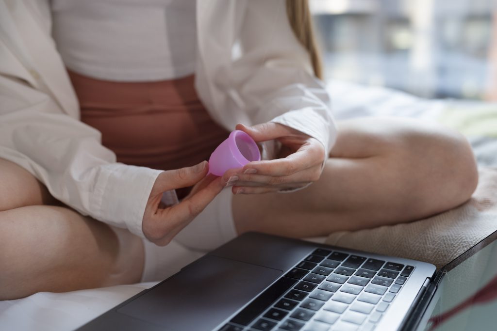 Uma pessoa sentada na cama, segurando um copo menstrual rosa, com um laptop preto ao seu lado. Ela usa uma camisa branca e uma saia rosa.