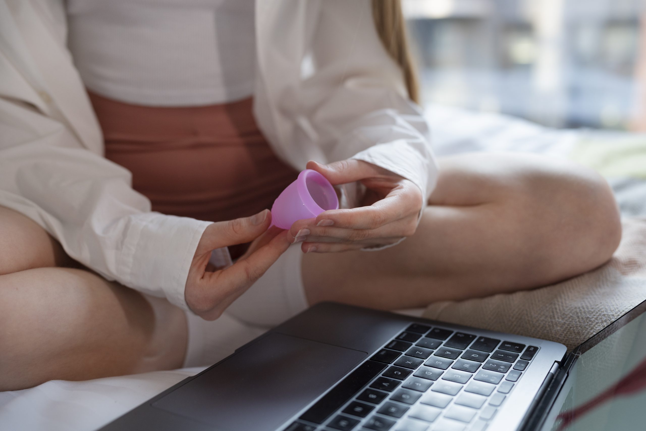 Uma pessoa sentada na cama, segurando um copo menstrual rosa, com um laptop preto ao seu lado. Ela usa uma camisa branca e uma saia rosa.