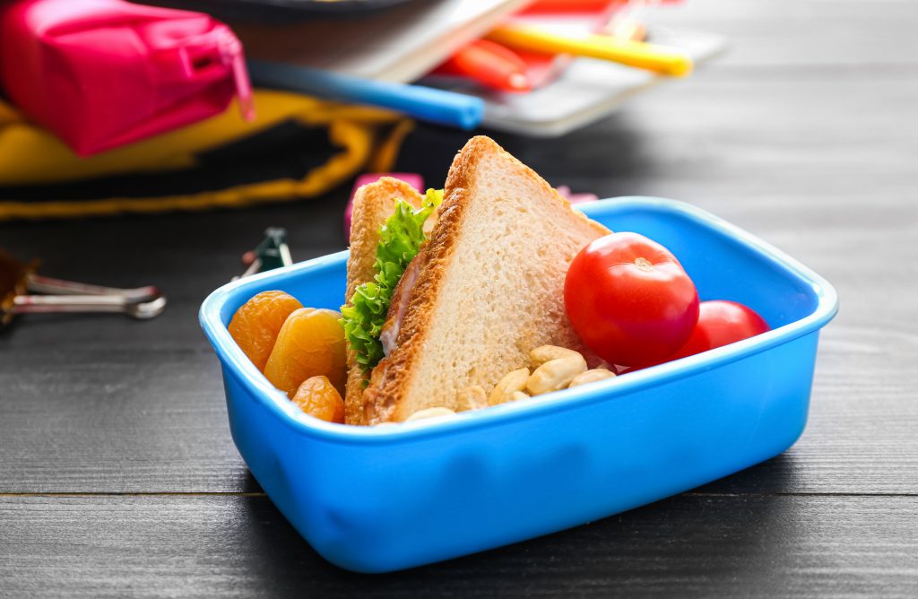 Lancheira azul com alimentação saudável: um sanduíche, tomates, damascos e nozes, sobre uma mesa escura.