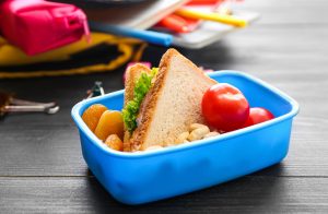 Lancheira azul com alimentação saudável: um sanduíche, tomates, damascos e nozes, sobre uma mesa escura.