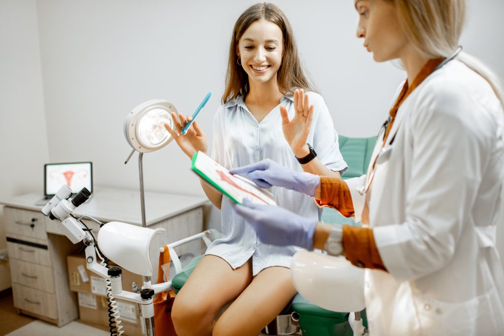 Uma paciente sorridente sentada em uma cadeira de exame em um consultório médico, enquanto uma profissional de saúde, com luvas, mostra uma folha com informações. Ambas estão em um ambiente clínico, com equipamentos médicos visíveis ao fundo.