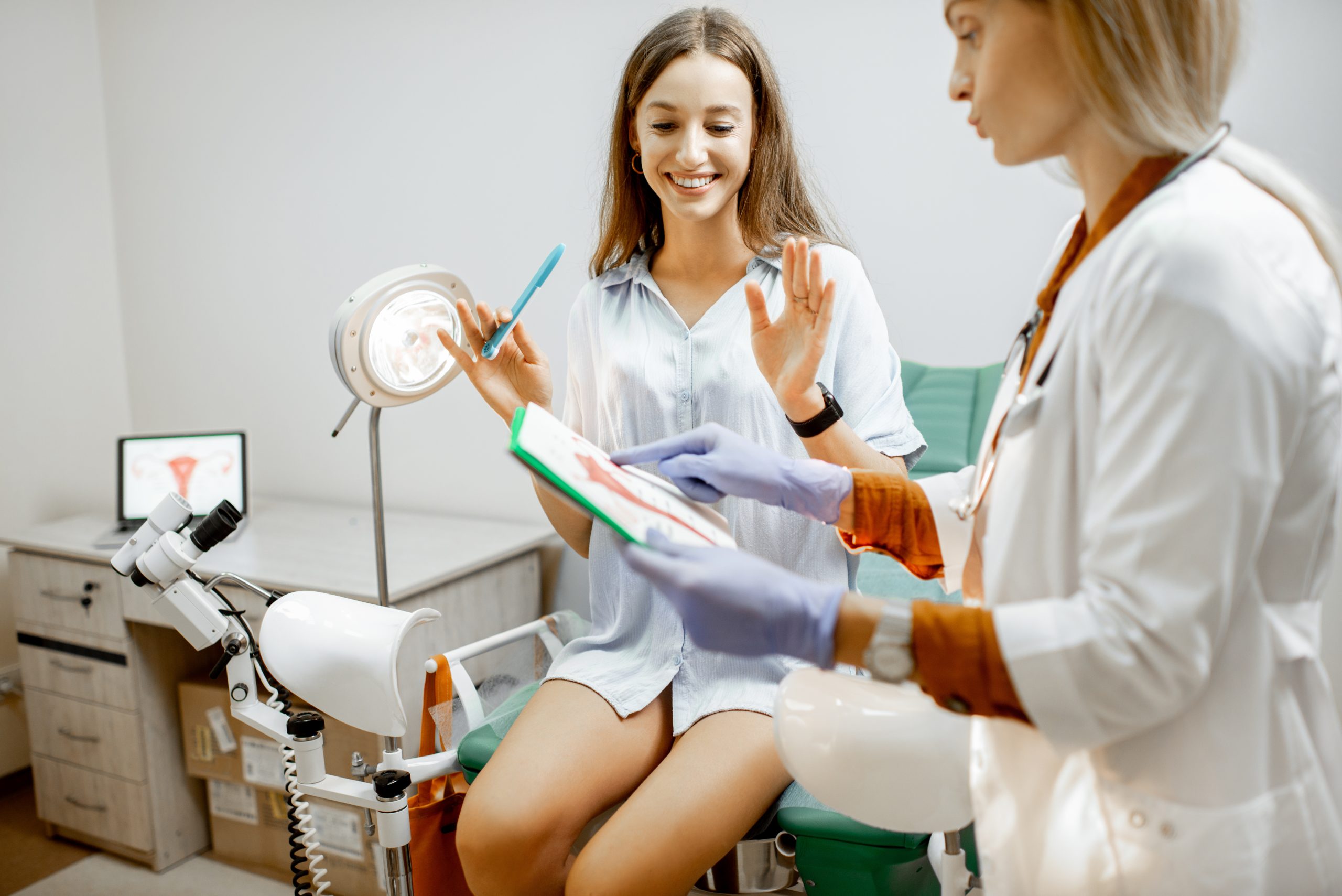 Uma paciente sorridente sentada em uma cadeira de exame em um consultório médico, enquanto uma profissional de saúde, com luvas, mostra uma folha com informações. Ambas estão em um ambiente clínico, com equipamentos médicos visíveis ao fundo.