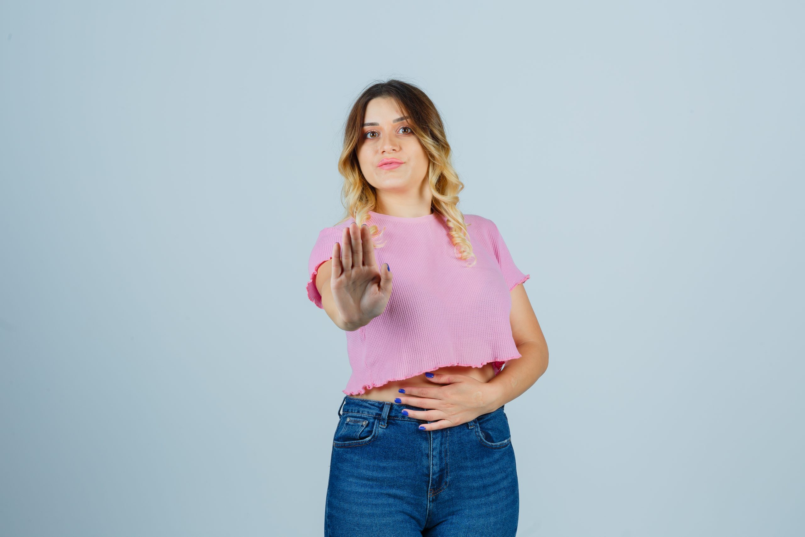 Mulher com blusa rosa e calça jeans, fazendo um gesto de "pare" com a mão enquanto a outra mão repousa sobre o abdômen. Fundo neutro.