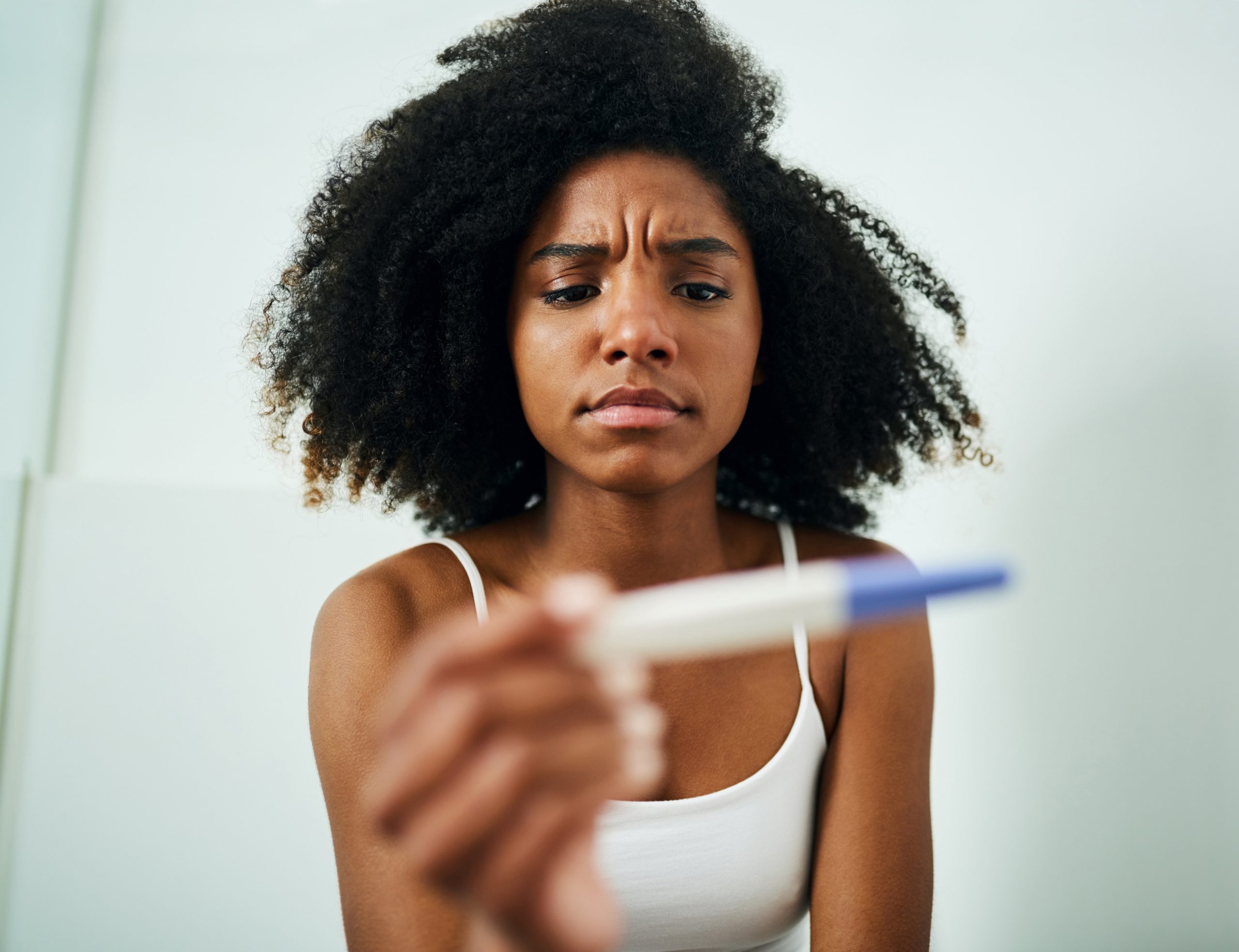 Mulher com cabelo cacheado, olhando para um teste de gravidez, com expressão preocupada. Ela tem infertilidade e está vestindo uma regata branca.