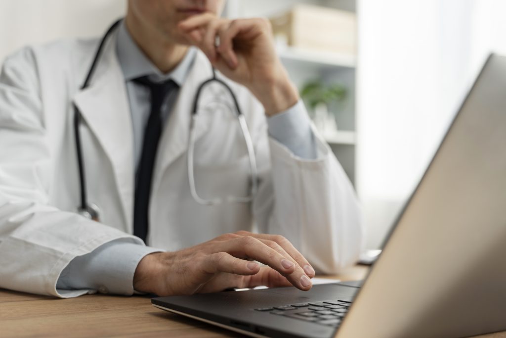 Um médico em um jaleco branco, com um estetoscópio no pescoço, digitando em um laptop, enquanto parece pensativo. O ambiente inclui prateleiras ao fundo com plantas e objetos decorativos.