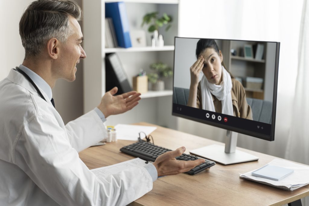 Um médico conversando com uma paciente por videoconferência, mostrando uma expressão de preocupação, enquanto a paciente parece estar estressada e tocando a testa. O ambiente é um consultório com uma mesa de madeira e uma tela de computador exibindo a imagem da paciente.