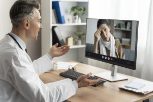 Um médico conversando com uma paciente por videoconferência, mostrando uma expressão de preocupação, enquanto a paciente parece estar estressada e tocando a testa. O ambiente é um consultório com uma mesa de madeira e uma tela de computador exibindo a imagem da paciente.