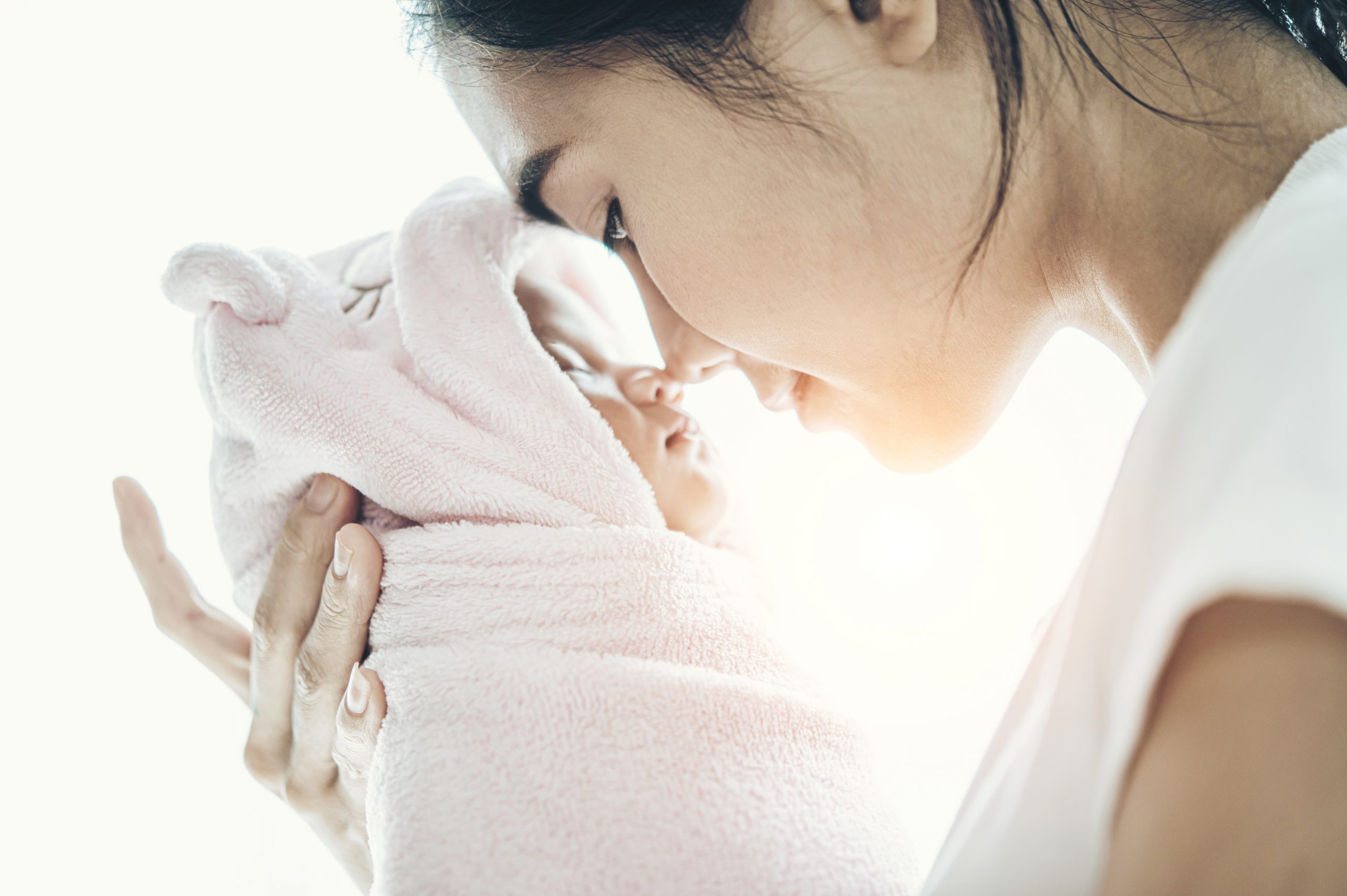 Uma mãe acaricia o rosto de um bebê enrolado em uma toalha rosa, enquanto ambos se aproximam em um gesto carinhoso. O fundo é claro, com luz suave.
