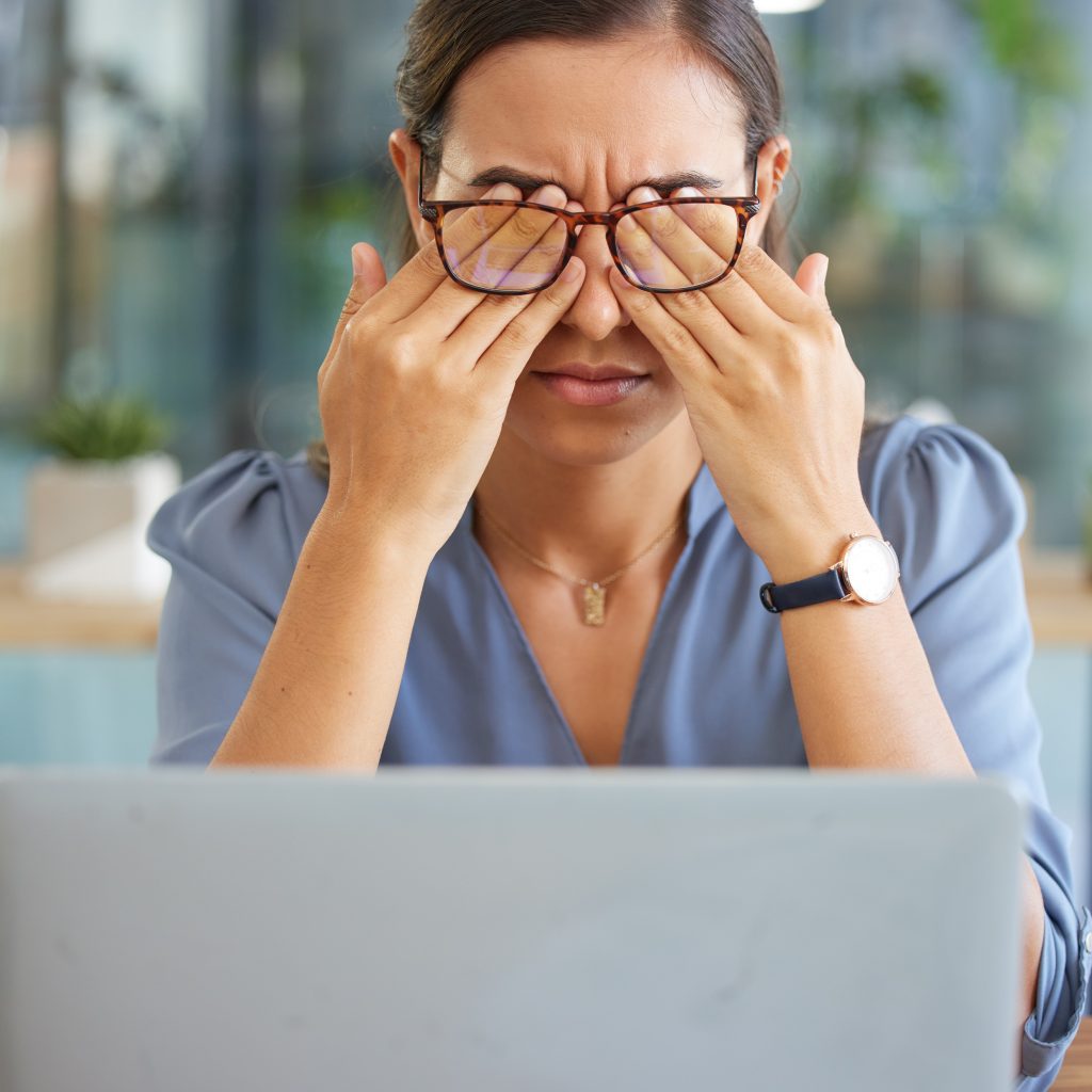 Mulher com blusa azul, usando óculos, esfregando os olhos com as mãos enquanto olha para um laptop em um ambiente de escritório iluminado.