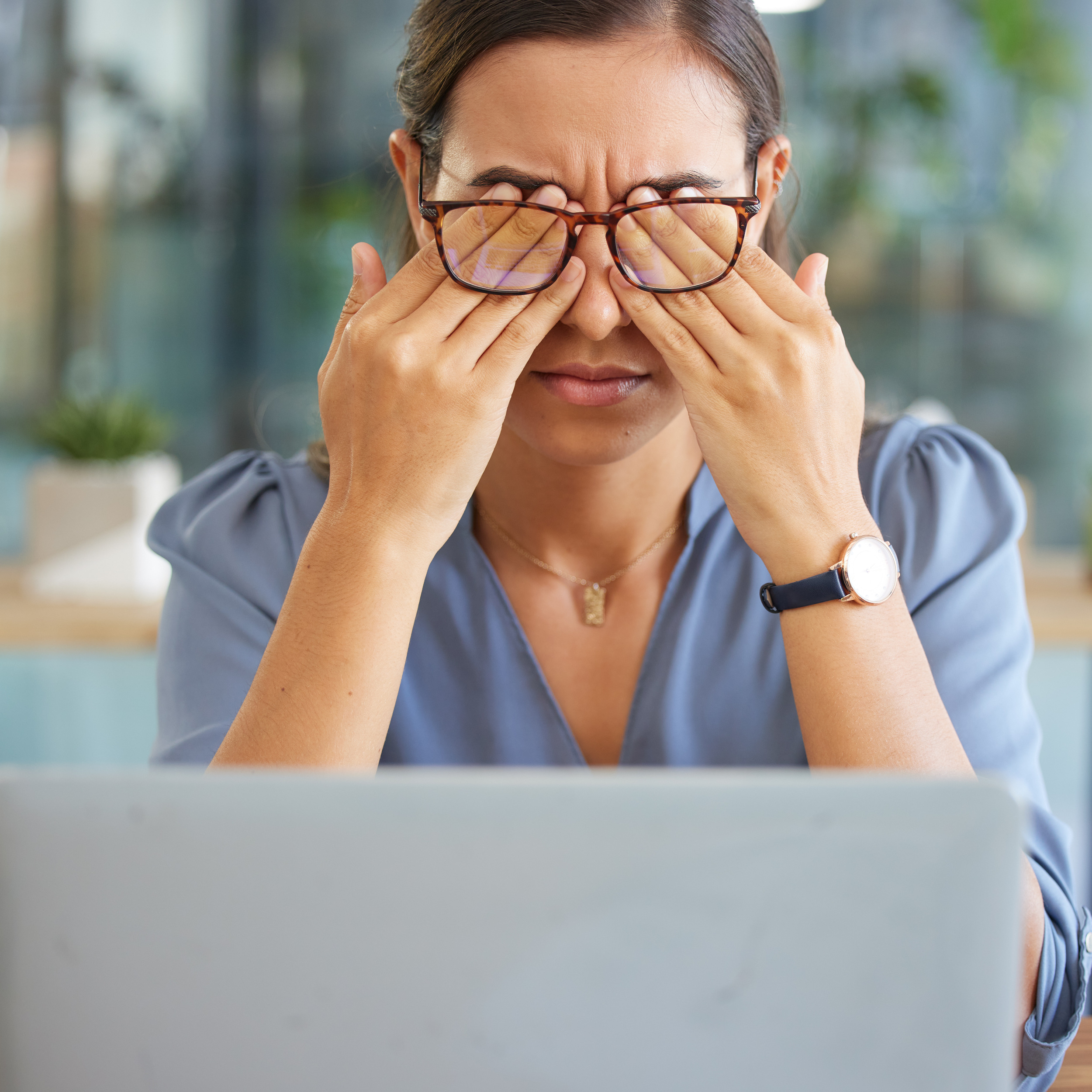 Mulher com blusa azul, usando óculos, esfregando os olhos com as mãos enquanto olha para um laptop em um ambiente de escritório iluminado.
