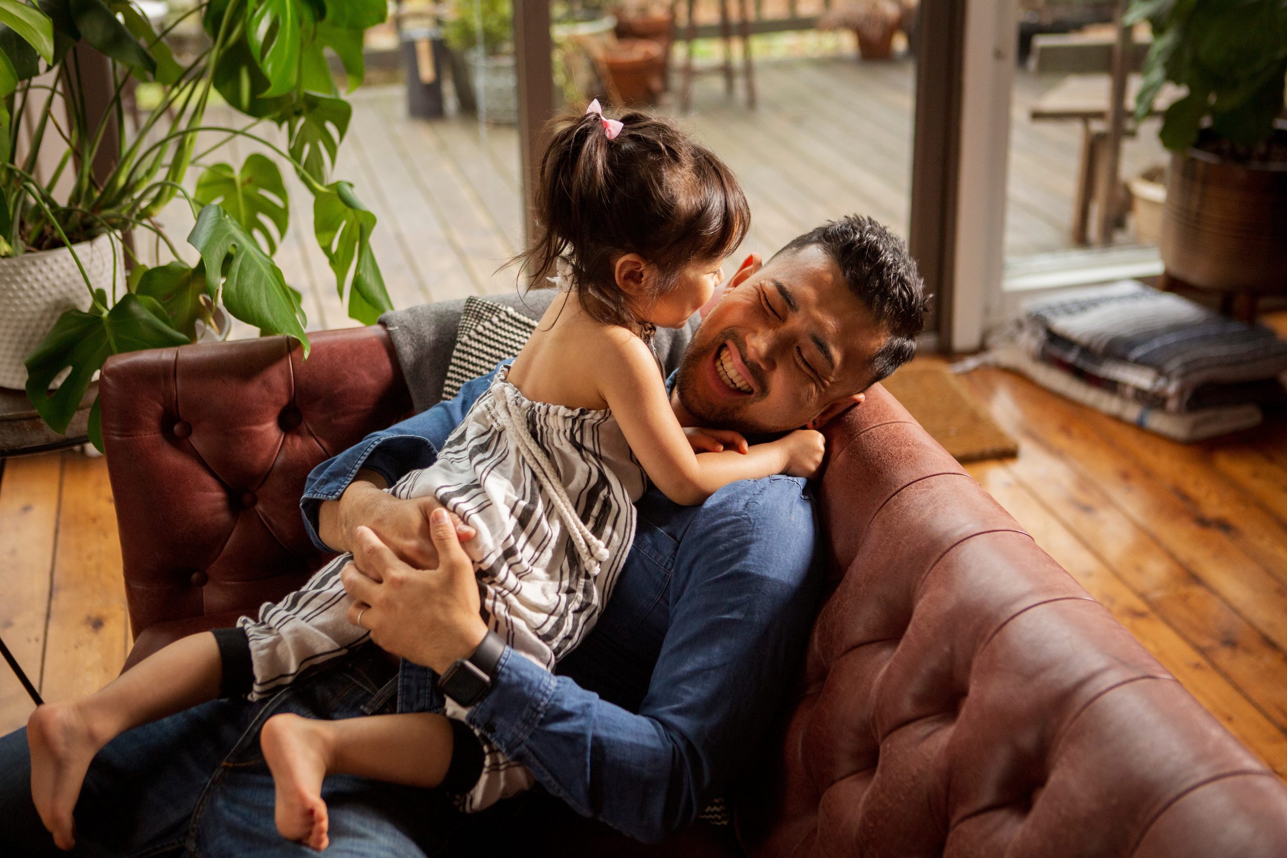 Um pai sorridente sentado em um sofá de couro marrom, segurando sua filha pequena, que está vestida com um macacão listrado, enquanto ela sorri e se inclina para ele. O ambiente é iluminado por luz natural, com plantas ao fundo e piso de madeira visível.
