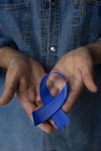 Uma fita azul em forma de laço é segurada com duas mãos, enquanto a pessoa usa uma camisa de jeans.