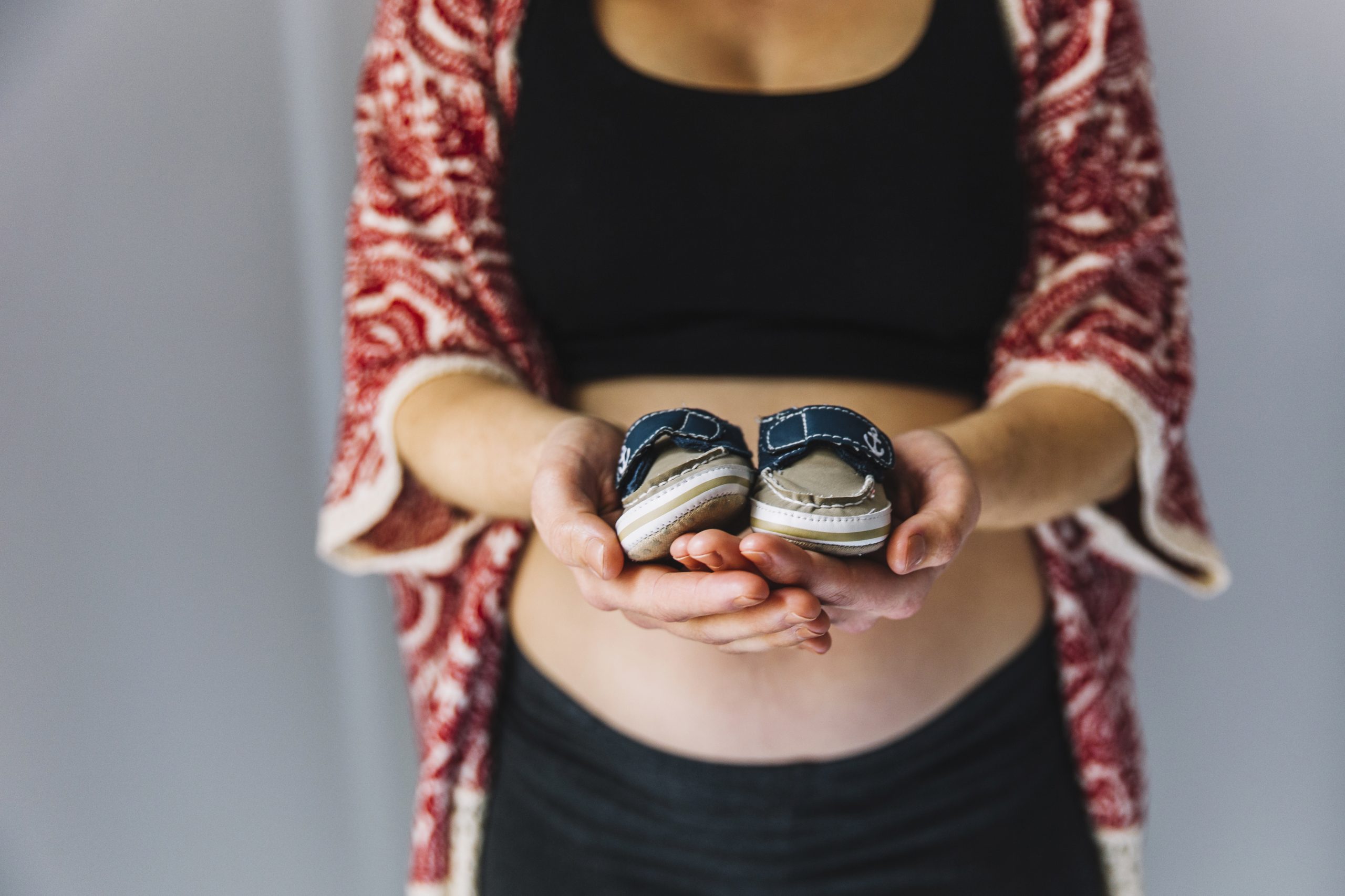 Mulher segurando um par de sapatos infantis em suas mãos, vestindo uma blusa preta e um casaquinho com padrões vermelhos; sua barriga está visível, sugerindo gravidez.