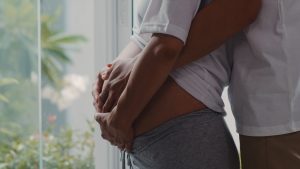 young asian pregnant couple hug holding belly talking with their child mom dad feeling happy smiling peaceful while take care baby pregnancy near window living room home Central de Consultas | Clínica Popular
