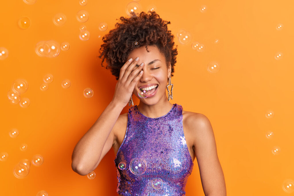 Dor de cabeça depois do Carnaval: quando pode ser só rotina e quando investigar 1 positive afro american woman covers face with hand smiles broadly has perfect white teeth dressed stylish clothing isolated orange wall with soap bubbles around Central de Consultas | Clínica Popular
