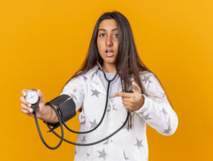 scared young ill girl measuring her own pressure with sphygmomanometer isolated yellow background Central de Consultas | Clínica Popular