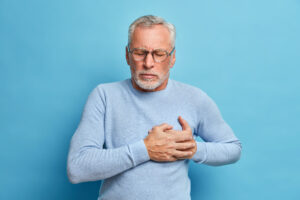 senior man spectacles presses hand chest has heart attack suffers from unbearable pain closes eyes wears optical glasses poses against blue wall Central de Consultas | Clínica Popular